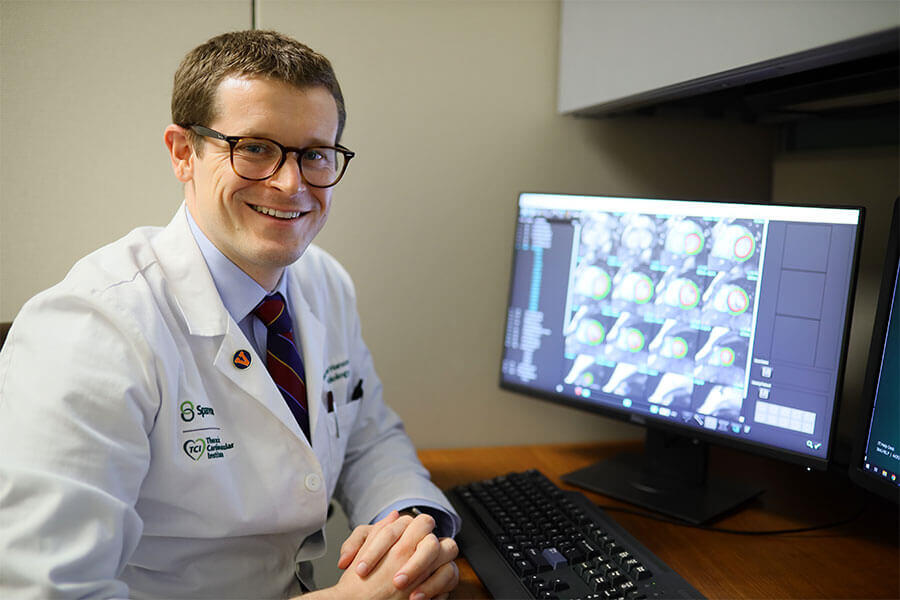 A smiling cardiologist in a white lab coat sitting next to a computer monitor displaying medical imaging scans.